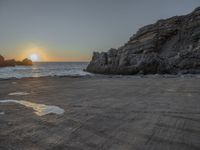 a lone motorcycle is parked near a rock formation on the beach at sunset, with water and ocean in the background