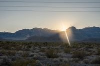 the sun is setting behind the mountains with bushes in foreground and power lines above