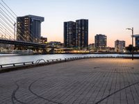 a man on a bike riding next to a river at sunset with the city lights of city buildings