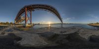 a bridge spans over the water near a shoreline at sunset time with a bright reflection of the sun coming out behind