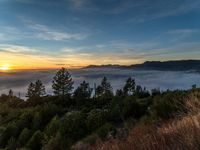 the sun setting over the clouds over trees in the mountains near an area with hills and small pine trees