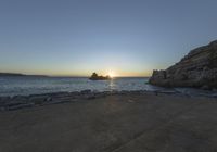 a beach near the ocean is seen at sunset with sun setting over the ocean and rocky shore