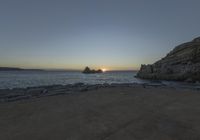 a beach near the ocean is seen at sunset with sun setting over the ocean and rocky shore