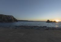 the sun is setting in front of a rocky shoreline and the ocean is below the rocks