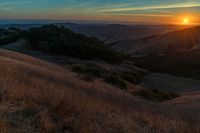 there is a sunset in the horizon on a field with grassy land below the mountains