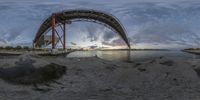 a view of the sunset under a large metal bridge over water and a sand beach