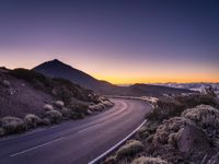 an empty winding road on a mountain with the sun set above the mountains in the background