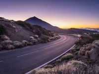 an empty winding road on a mountain with the sun set above the mountains in the background