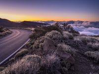 an empty winding road on a mountain with the sun set above the mountains in the background