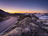 an empty winding road on a mountain with the sun set above the mountains in the background