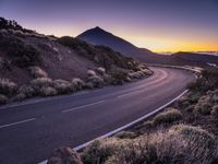 an empty winding road on a mountain with the sun set above the mountains in the background