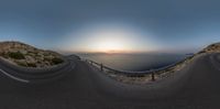 a picture of the sun setting over the ocean and road from the street level looking down to the ocean