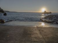 the sun is setting over the water at a coast with rocks in it, and a boat is sailing in the background