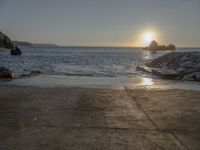 the sun is setting over the water at a coast with rocks in it, and a boat is sailing in the background