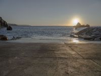 the sun is setting over the water at a coast with rocks in it, and a boat is sailing in the background