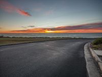 the sun sets over the ocean on an empty road near the water and grass and bushes