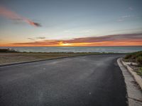 the sun sets over the ocean on an empty road near the water and grass and bushes