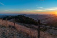 a sunset in the distance over a valley with a fence and tree to the foreground