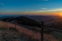 a sunset in the distance over a valley with a fence and tree to the foreground