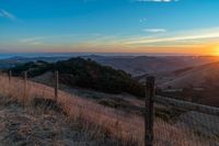 a sunset in the distance over a valley with a fence and tree to the foreground