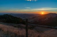 a sunset in the distance over a valley with a fence and tree to the foreground