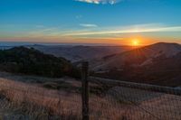 a sunset in the distance over a valley with a fence and tree to the foreground