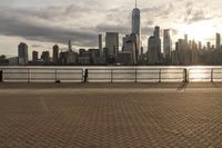a person on the boardwalk near a fence overlooking a river and city skyline at sunset