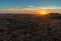 the sunset is reflecting off the mountain range above the grass field on a hill that looks like a hillside