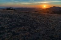 the sunset is reflecting off the mountain range above the grass field on a hill that looks like a hillside