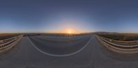 a wide angle view of the road near a fenced off area at sunset with the sun setting in the distance