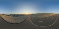 a panoramic view shows the road and sand at sunset with the sun setting