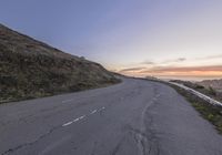 an empty empty road at sunset with traffic going by on the side of it near a cliff