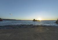 the sun is setting on the horizon in a rocky beach area, with some rocks and the water behind the shore