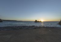 the sun is setting on the horizon in a rocky beach area, with some rocks and the water behind the shore