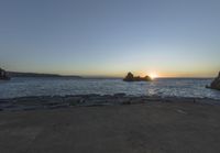 the sun is setting on the horizon in a rocky beach area, with some rocks and the water behind the shore