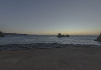 the sun is setting on the horizon in a rocky beach area, with some rocks and the water behind the shore