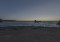 the sun is setting on the horizon in a rocky beach area, with some rocks and the water behind the shore