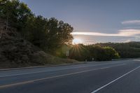 a car that is parked on the side of a road at sunset in a highway with the sun breaking through the trees