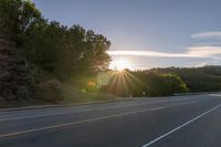 a car that is parked on the side of a road at sunset in a highway with the sun breaking through the trees