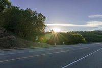 a car that is parked on the side of a road at sunset in a highway with the sun breaking through the trees