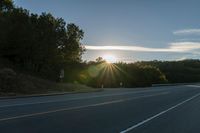 a car that is parked on the side of a road at sunset in a highway with the sun breaking through the trees