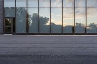 a sidewalk leading up to a glass walled building with trees in the background at sunset