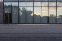 a sidewalk leading up to a glass walled building with trees in the background at sunset