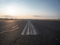 the sun is setting on a runway with a dotted path going to the left, at an airport