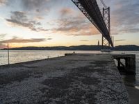 sunset view of the bay from beneath of a bridge over water with concrete pier and bench at the end of the pier