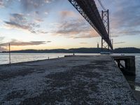 sunset view of the bay from beneath of a bridge over water with concrete pier and bench at the end of the pier