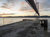 sunset view of the bay from beneath of a bridge over water with concrete pier and bench at the end of the pier