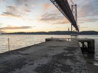 sunset view of the bay from beneath of a bridge over water with concrete pier and bench at the end of the pier