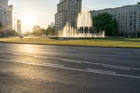 the sun is shining over a street and water feature fountain in the background of a big building