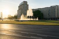 the sun is shining over a street and water feature fountain in the background of a big building